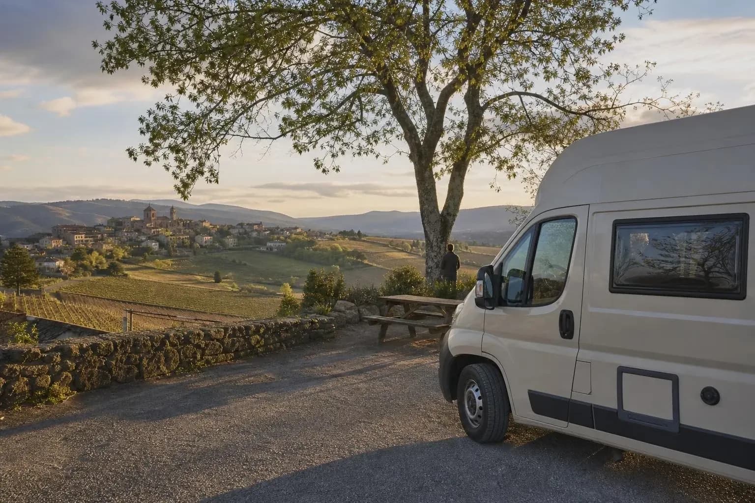Fourgon aménagé stationné sur un terre-plein gravillonné sous un grand arbre, près d’une table de pique-nique, face à un village perché et à des vignes au coucher du soleil