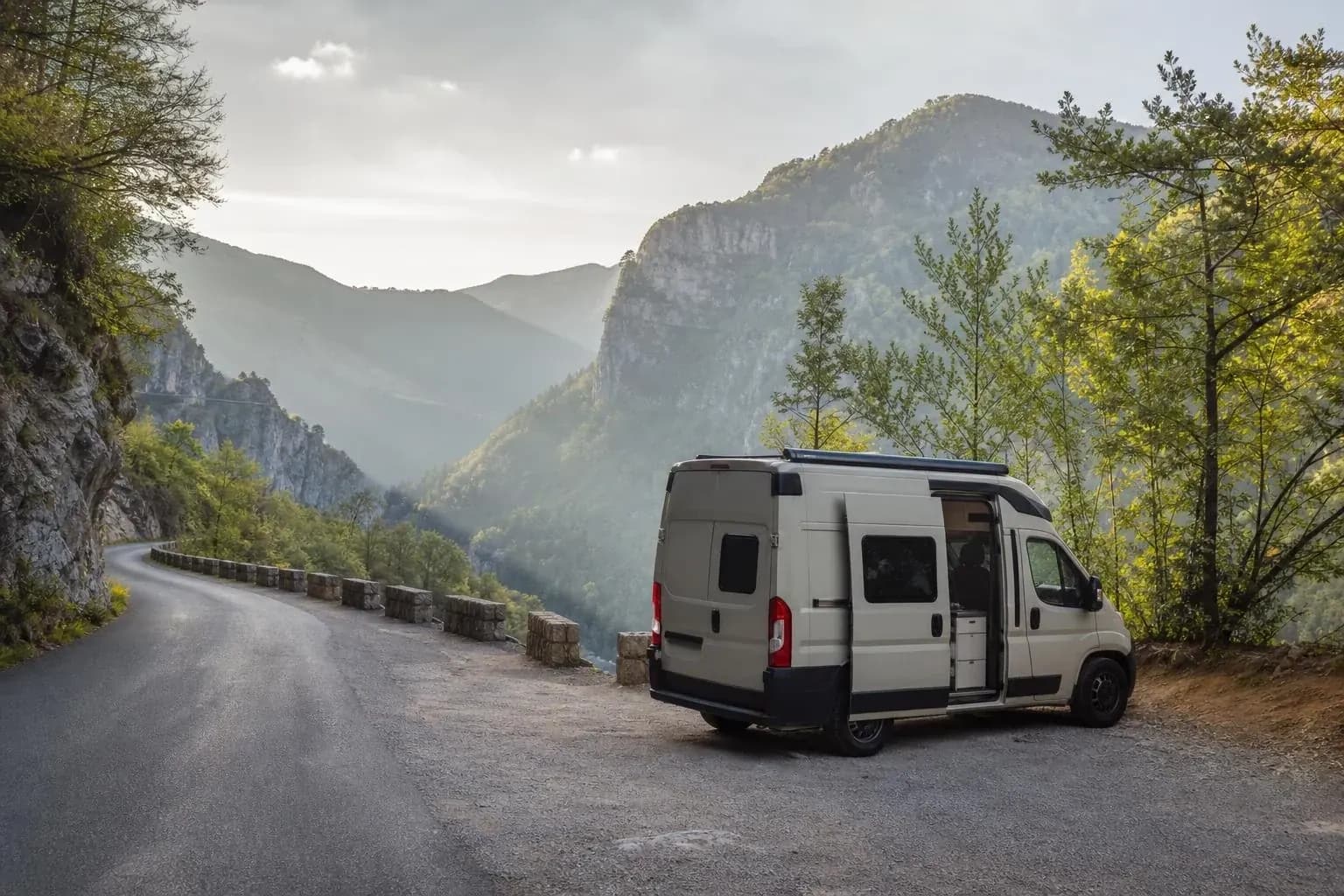 Fourgon aménagé porte latérale ouverte sur son coin cuisine, arrêté sur un élargissement gravillonné au bord d’une route de montagne, au-dessus d’une gorge boisée